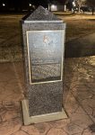 A granite monument with a plaque, set in a park during nighttime, surrounded by illuminated pathways and trees.