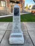 A granite monument with a carved image and inscriptions honoring Colonel Charles E. McKinney, located in an outdoor area with grass and buildings in the background.