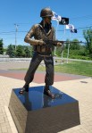 Bronze statue of a soldier in military uniform holding a rifle, set on a pedestal with flags in the background and a clear blue sky.