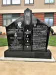 A black granite memorial monument featuring engraved portraits and text honoring individuals, with decorative elements and symbols, situated outdoors in front of a building.