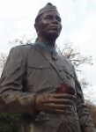 A bronze statue of a soldier in uniform, holding an apple, standing against a cloudy sky.