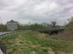 A scenic view of a roadside historical marker on a cloudy day, with grass and trees in the background.